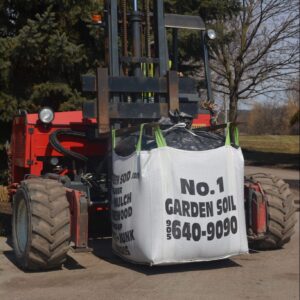 A red forklift carrying a large bag labeled "No. 1 Garden Soil" near a park with trees in the background.