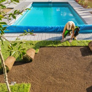 A person landscapes by laying sod near a swimming pool surrounded by decking and plants, creating a lush garden atmosphere under a clear sky.