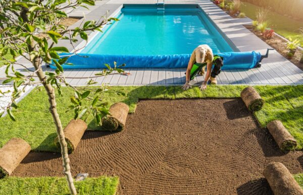 A person landscapes by laying sod near a swimming pool surrounded by decking and plants, creating a lush garden atmosphere under a clear sky.