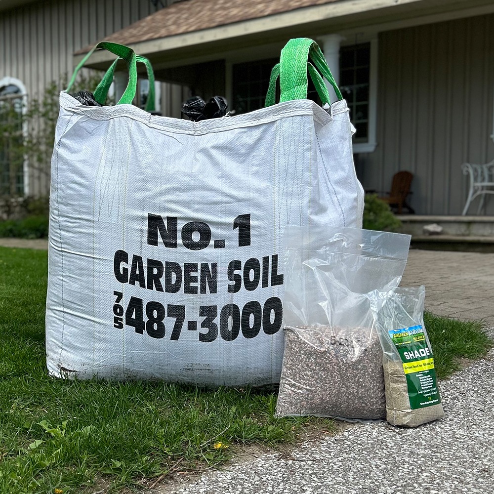 A large white garden soil bag and smaller bags of seeds sit on the grass near a house with a porch.