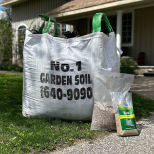 A large bag labeled "No. 1 Garden Soil" and a smaller bag of grass seed sit on a grassy lawn near a building's porch.