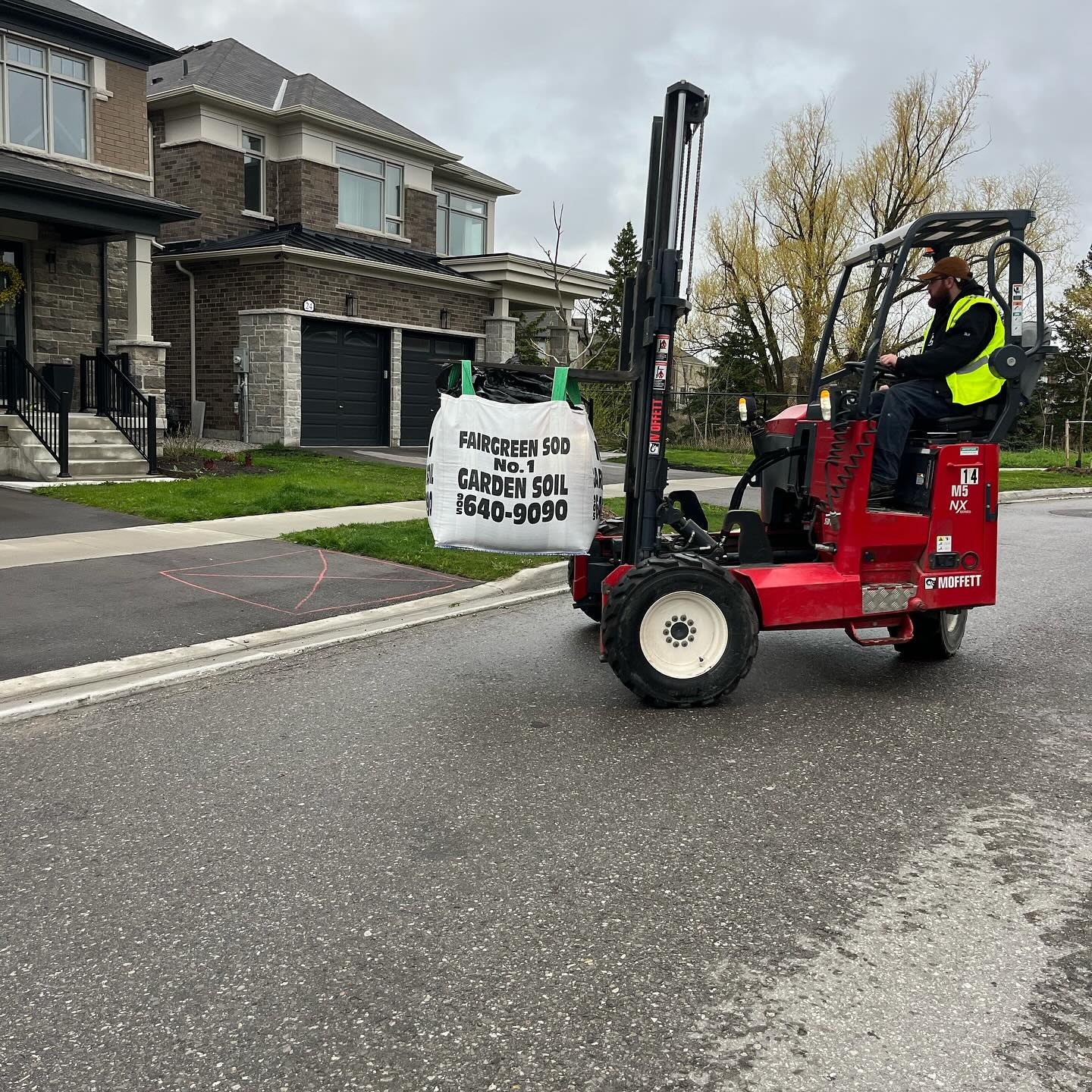 A person operates a forklift, transporting garden soil in a residential neighborhood. Two houses and a tree-lined street are visible near them.