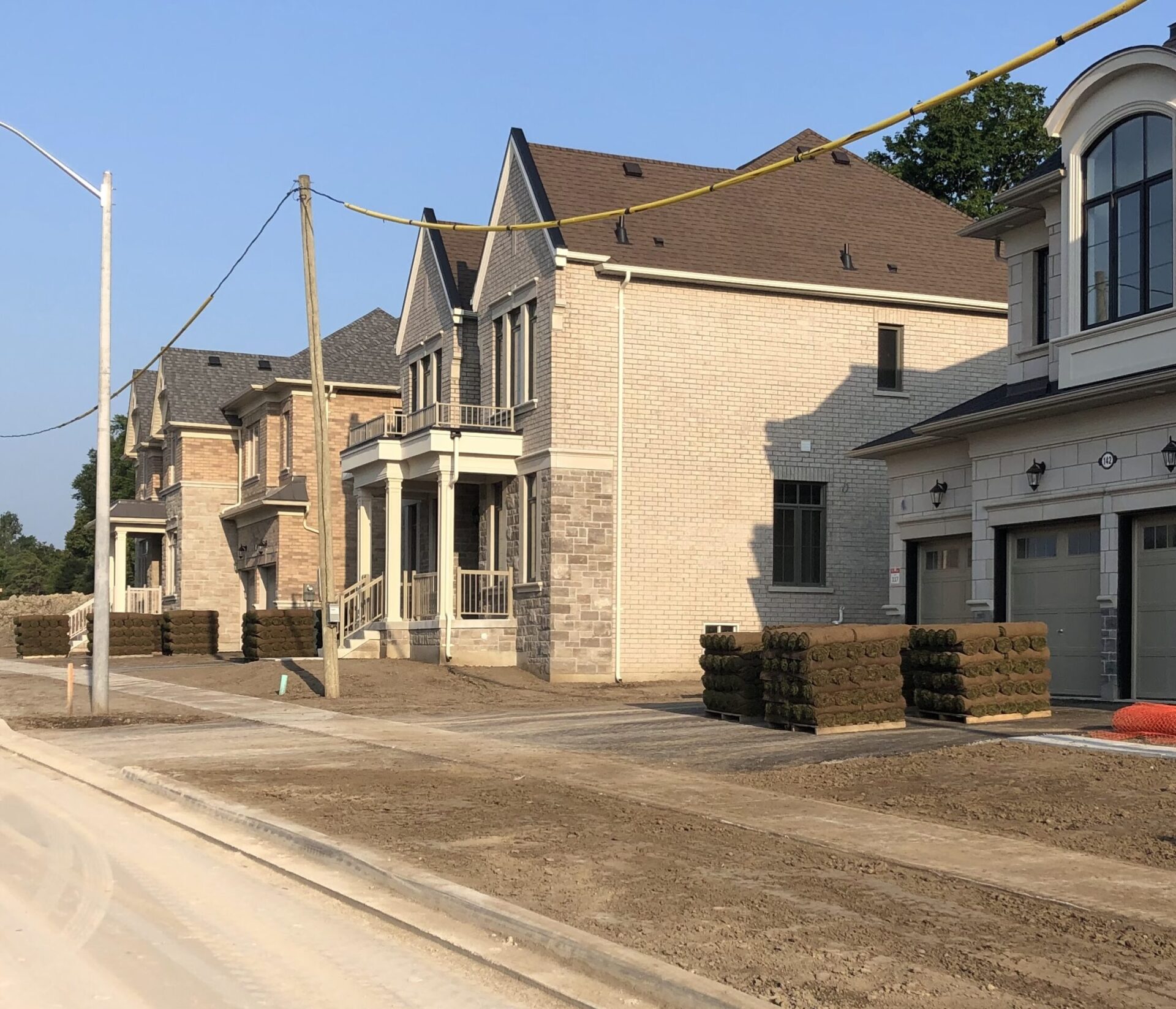 Newly constructed suburban houses with stacks of turf rolls along the sidewalk. No people or landmarks are visible. Clear, sunny day.