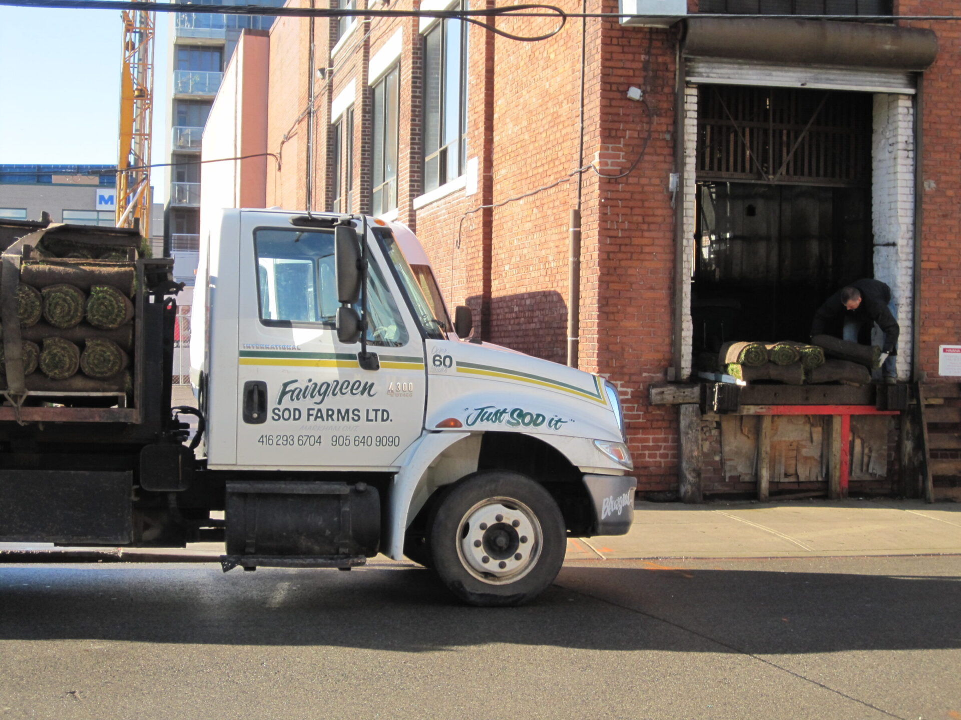 A truck labeled "Fairgreen Sod Farms Ltd." unloads rolled sod in front of a brick building, with a person assisting at a loading dock.