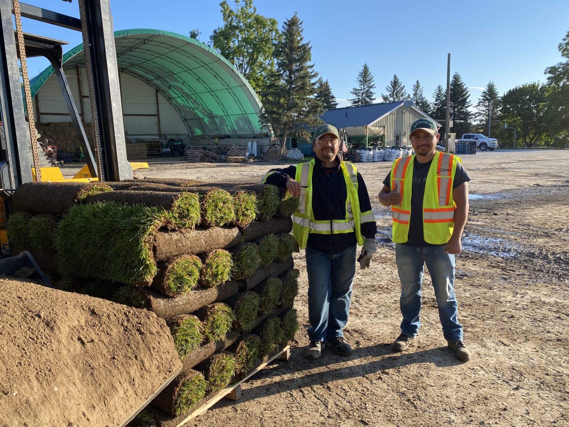 Two people in safety vests stand beside stacked sod rolls in a construction area, with a large storage tent in the background.