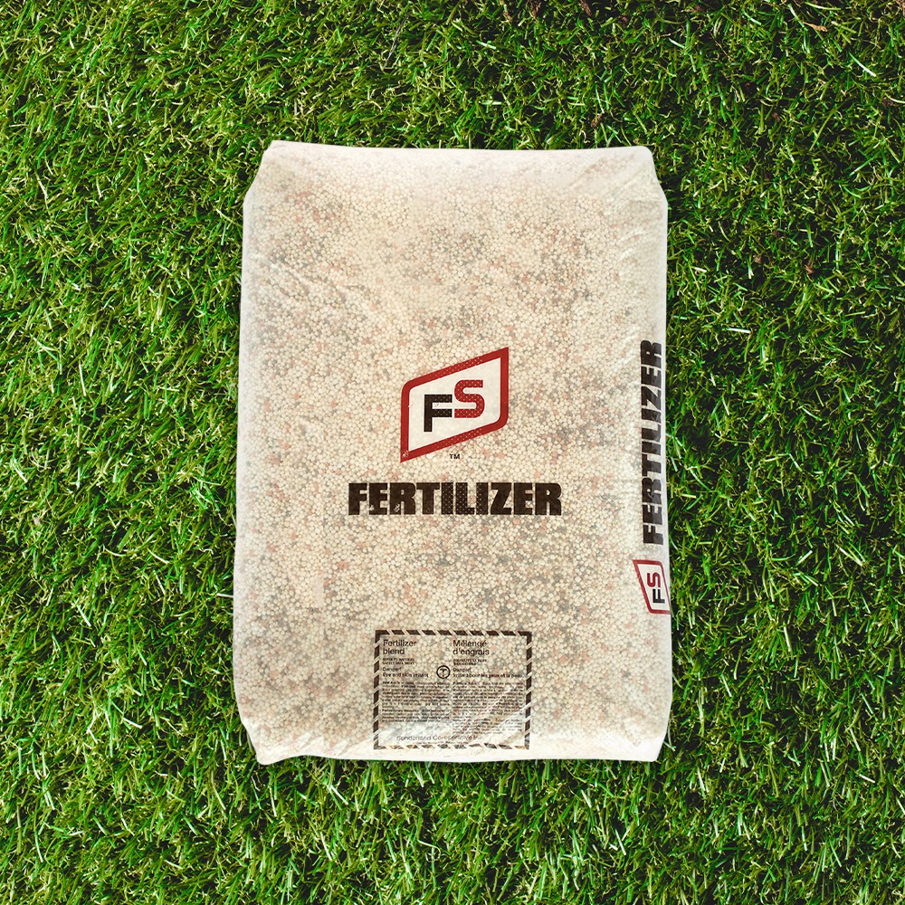 A bag labeled "FS Fertilizer" sits on green grass, filled with granular material. The product appears packaged for gardening or agricultural use.