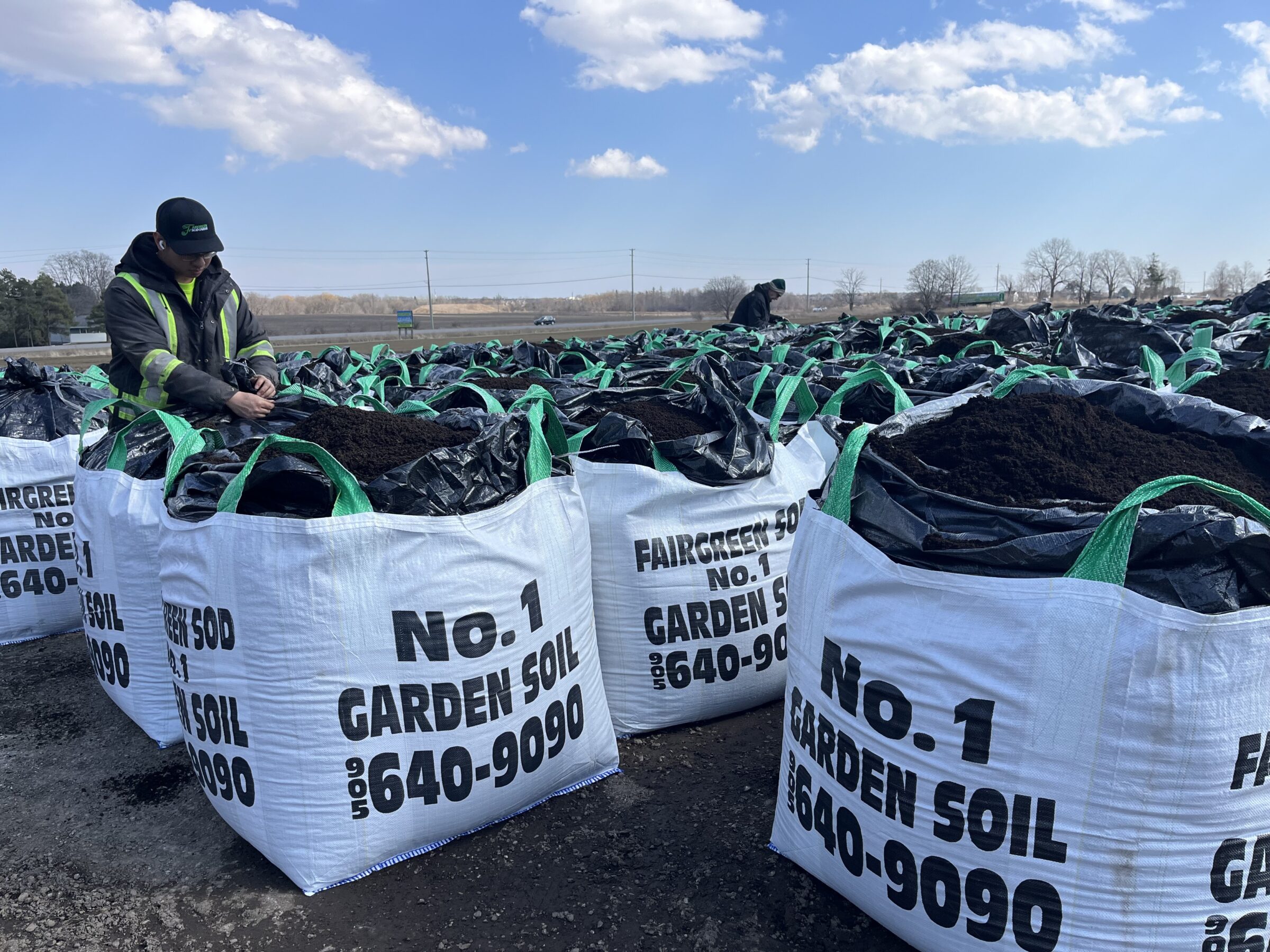 A person examines large bags of garden soil outdoors, surrounded by numerous similar bags under a partly cloudy sky.