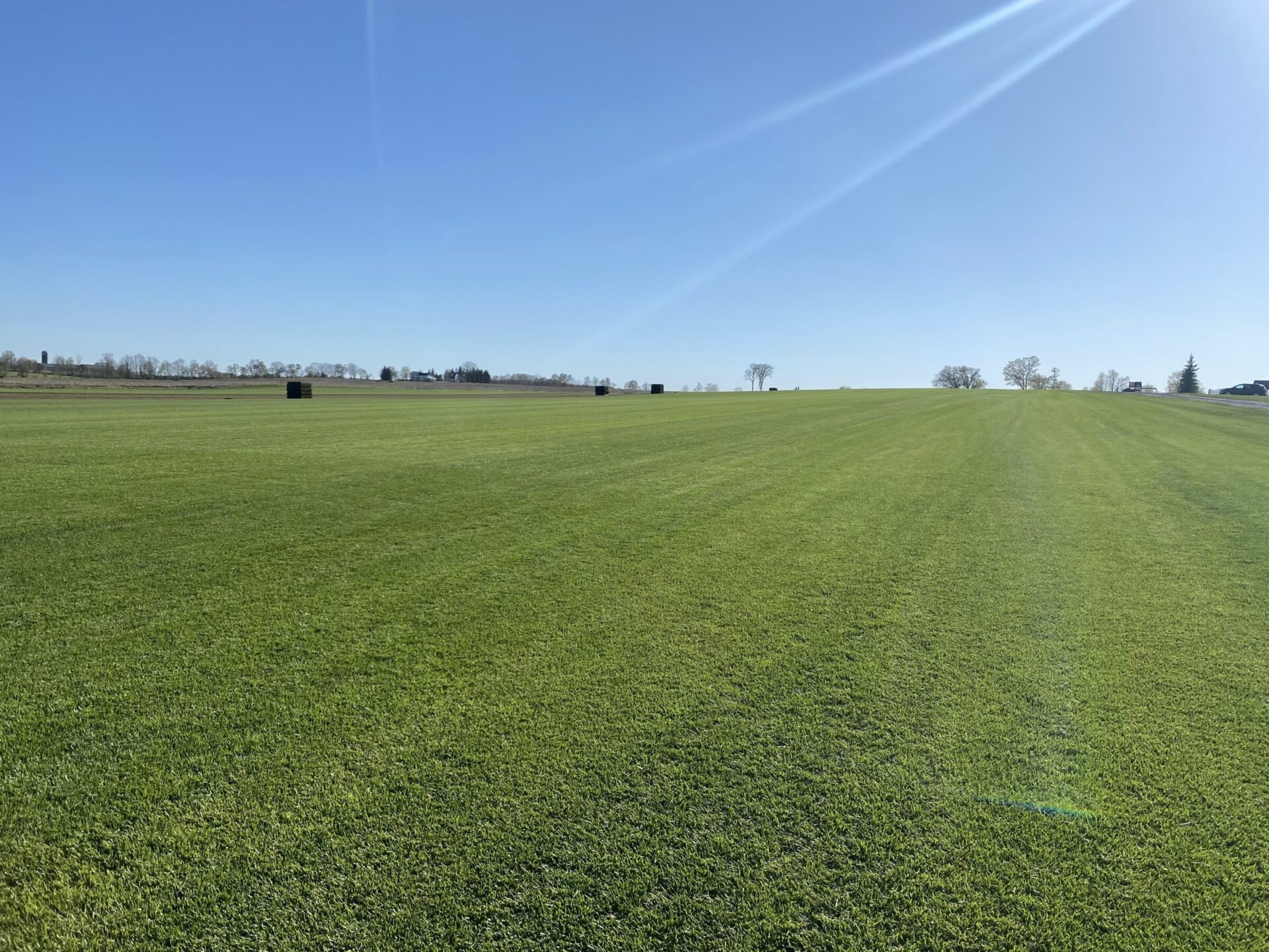 A vast, open field with neatly mowed grass under clear blue skies. Sparse trees and distant structures line the horizon.