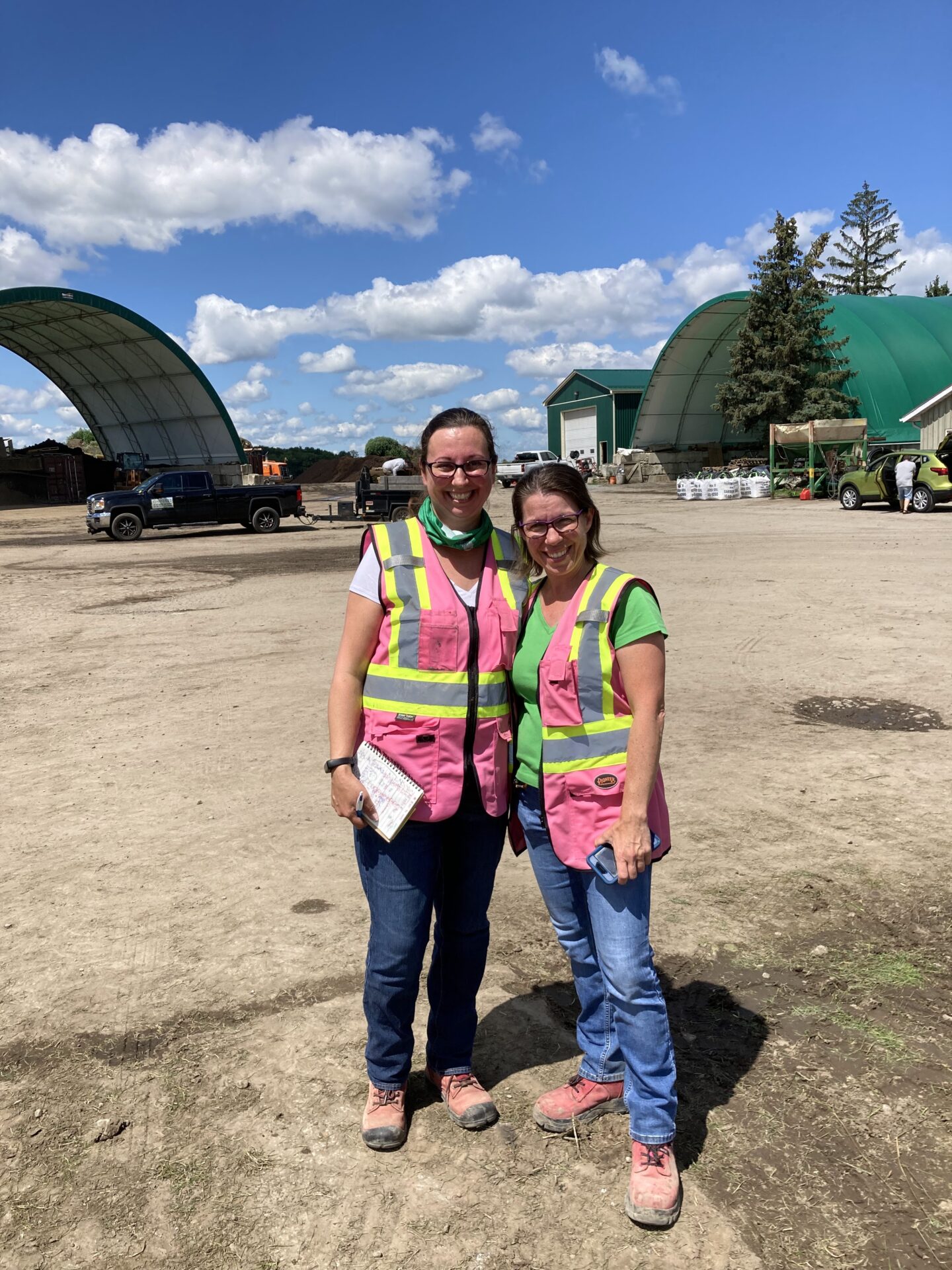 Two people wearing pink safety vests stand together at a construction site under a clear blue sky, with green-roofed structures in the background.