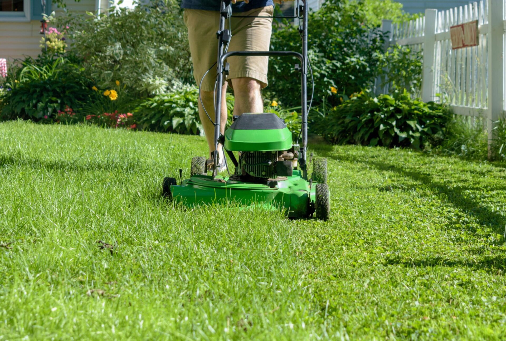 A person is mowing a lush green lawn with a push mower, surrounded by vibrant flowers and a white picket fence in a garden.