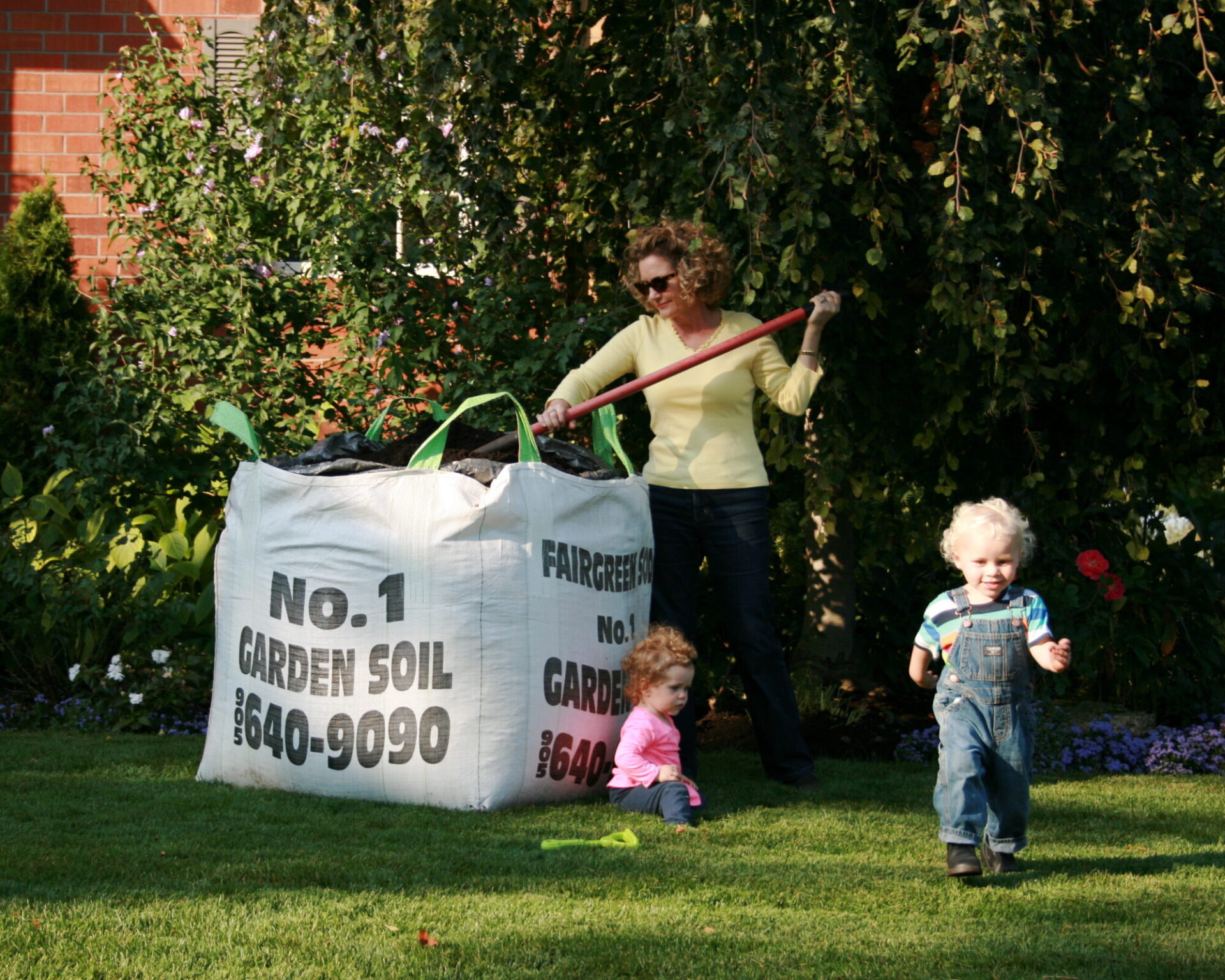 A person gardening with children nearby, beside a large bag of garden soil, in a lush green yard on a sunny day.