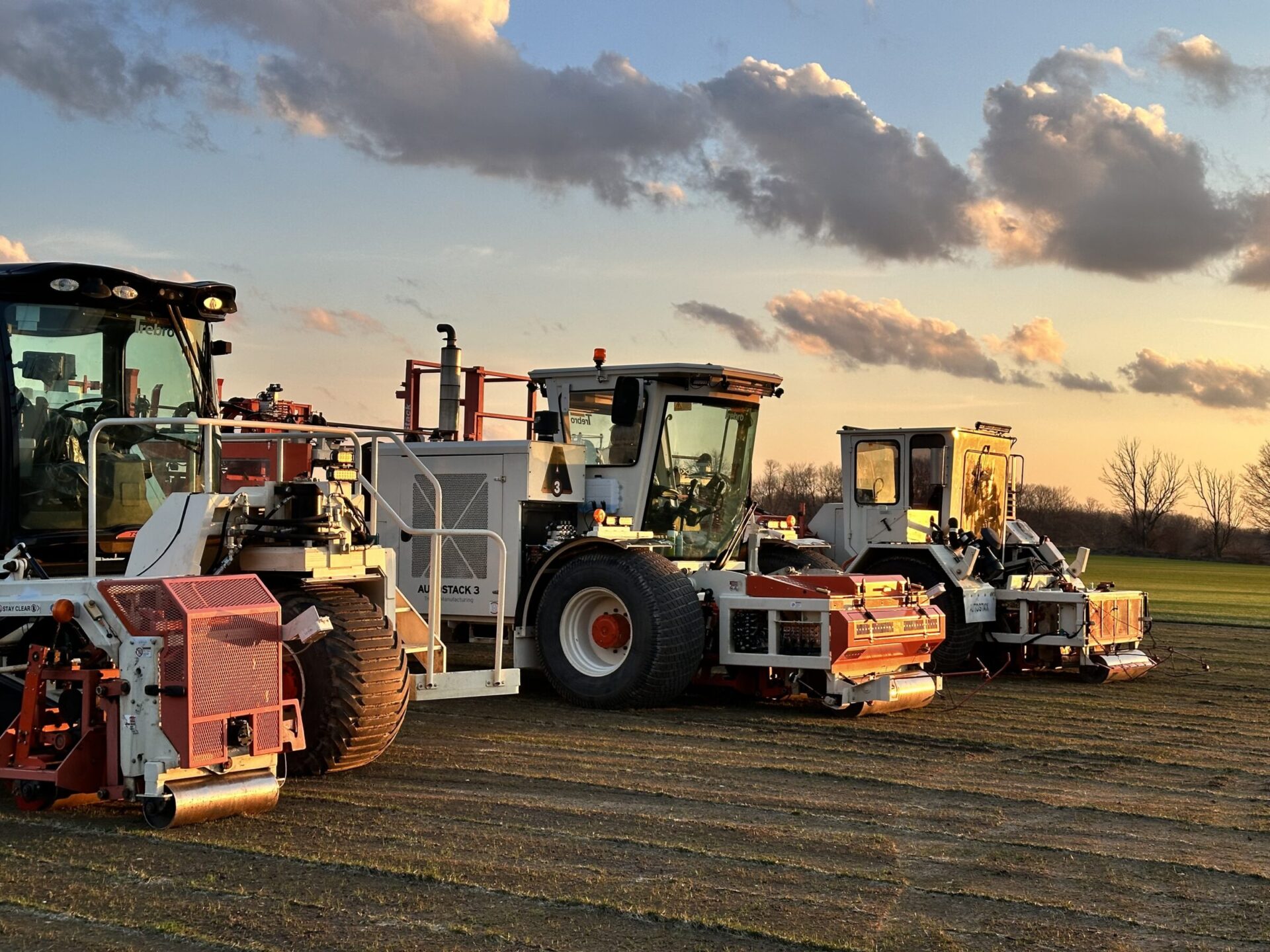 Several large agricultural machines are parked on a field at sunset, with a partly cloudy sky in the background. No people are present.