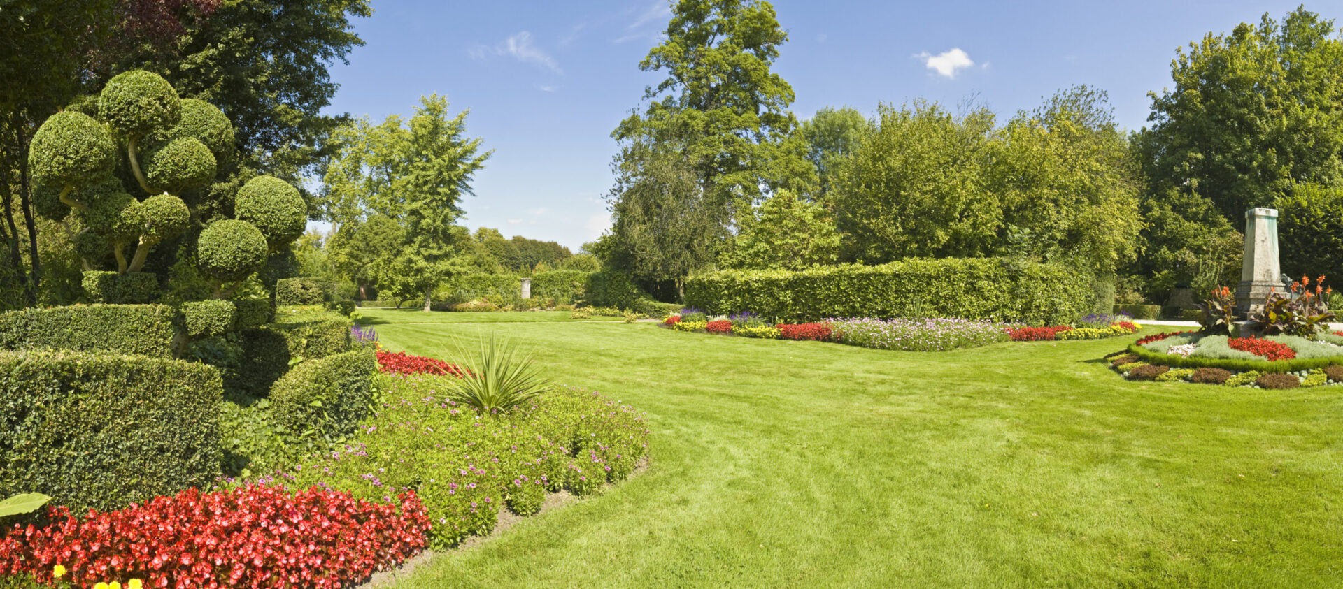 Lush garden with manicured hedges, vibrant flower beds, and a tall monument surrounded by trees under a clear blue sky.