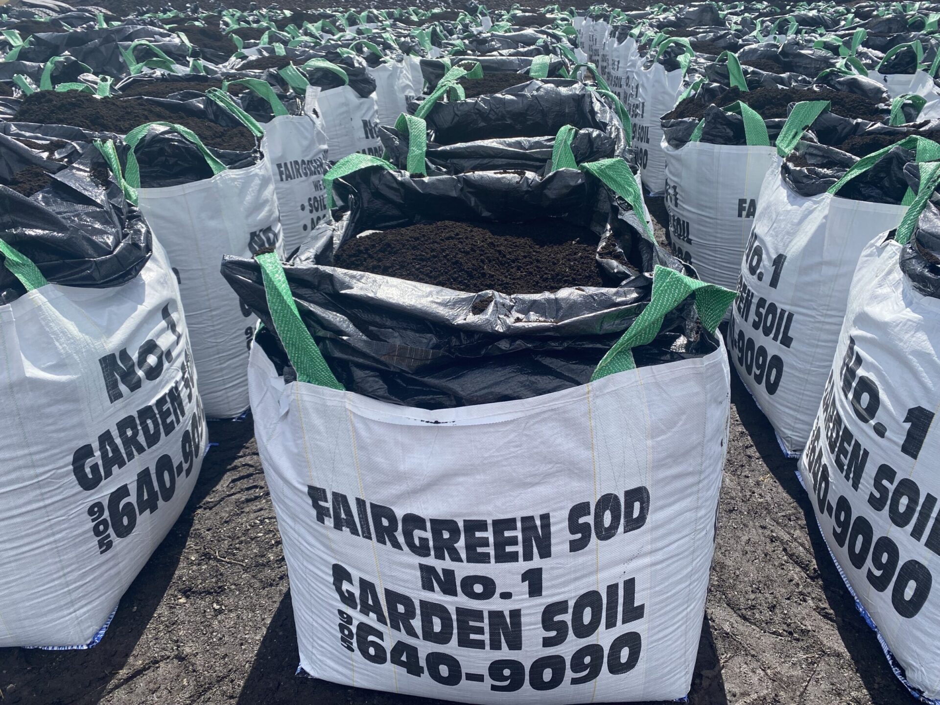 Large white bags filled with garden soil, labeled "Fairgreen Sod No. 1," are arranged in rows on a dirt surface.