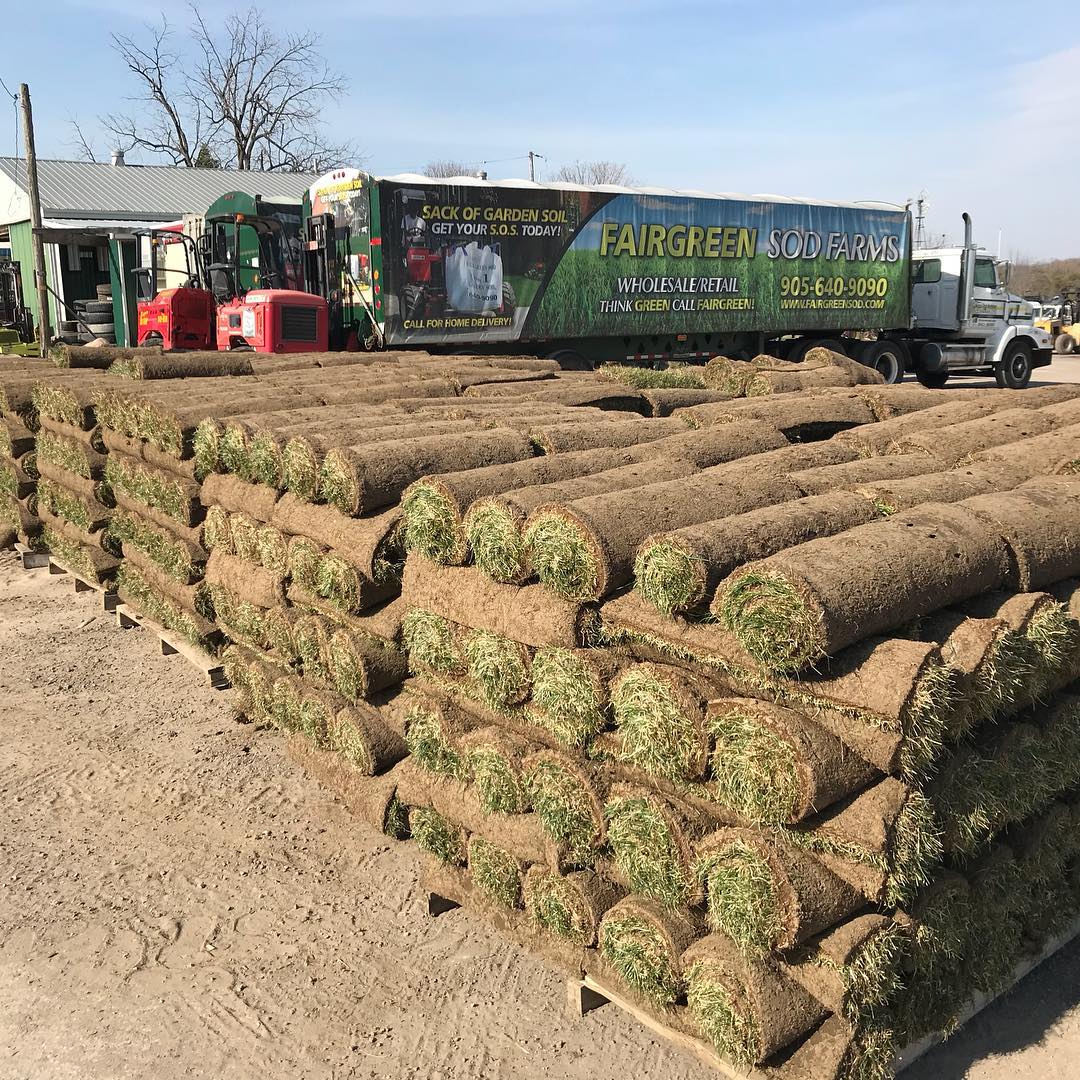 Stacks of rolled sod ready for delivery at Fairgreen Sod Farms. A truck and farming equipment are visible in the background.