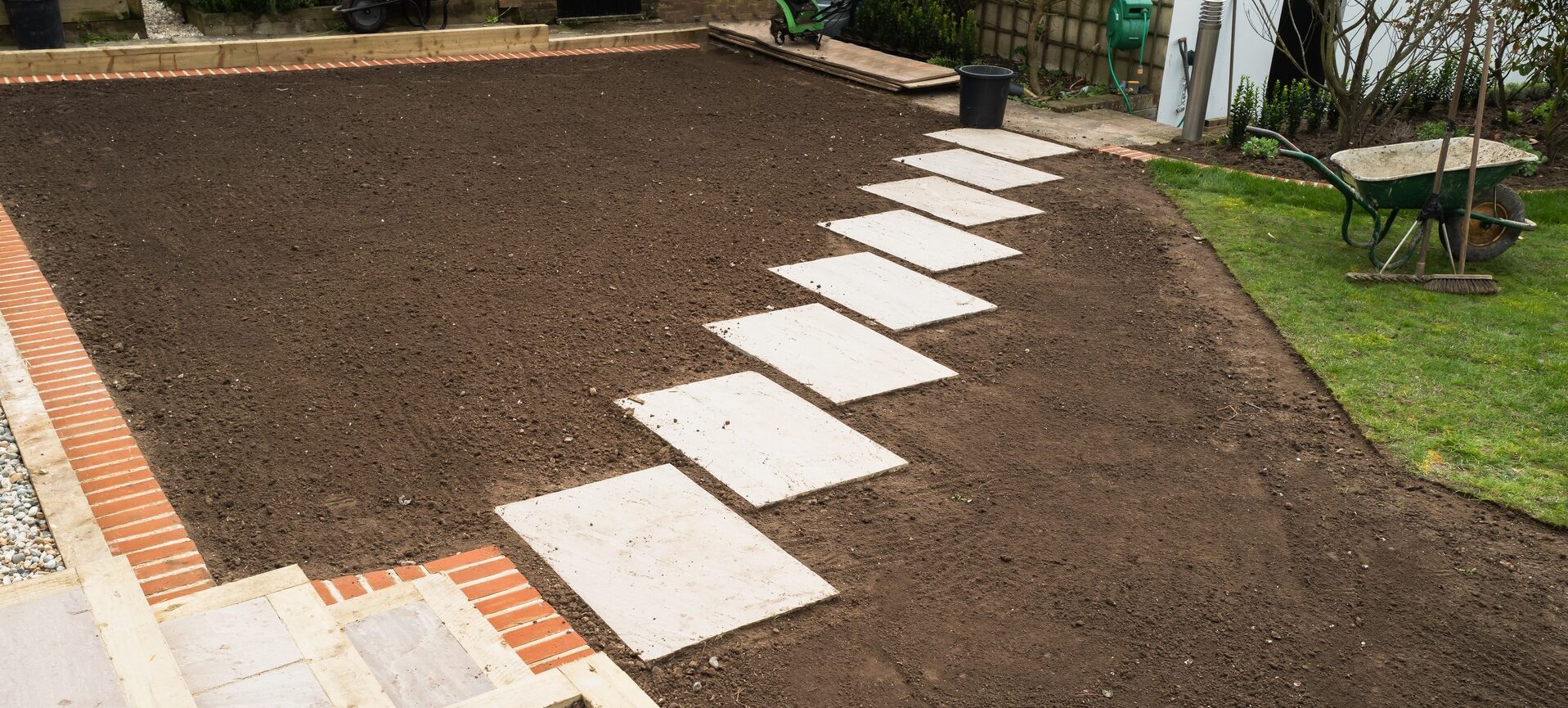 A garden with freshly turned soil, paving stones, and a wheelbarrow. Small plants are in a border next to a grass area.