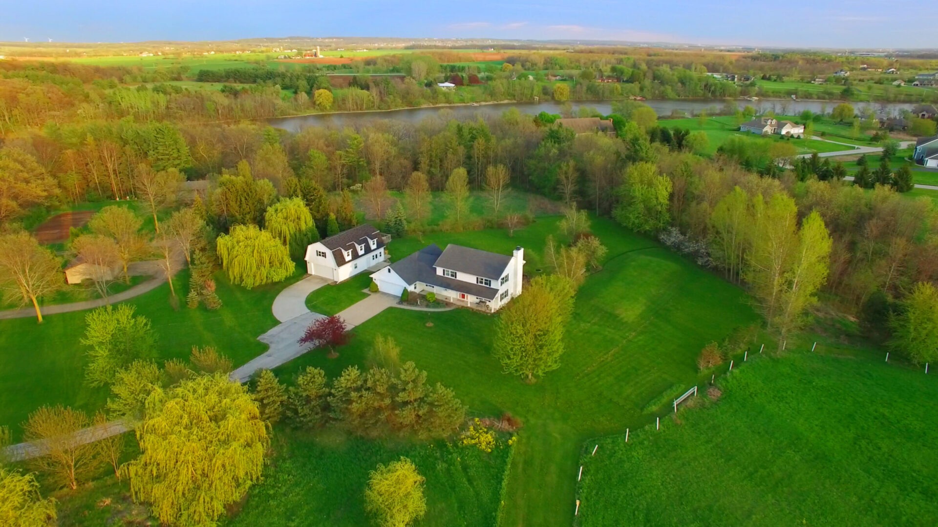 Aerial view of a countryside house surrounded by lush greenery, trees, and a winding driveway, with a peaceful pond nearby.