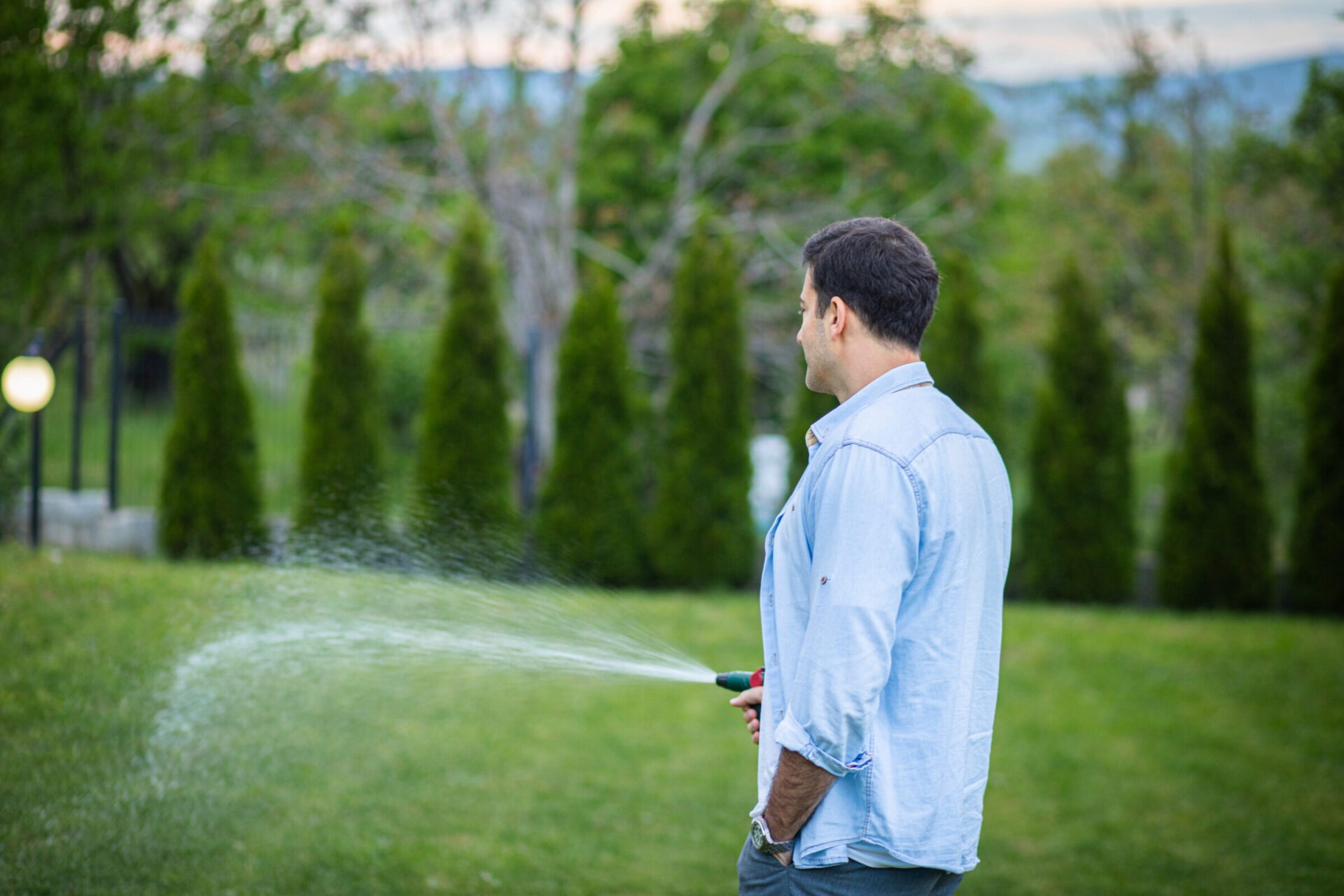 A person in a blue shirt waters green grass in a garden with trees, creating an arc of water from a hose.