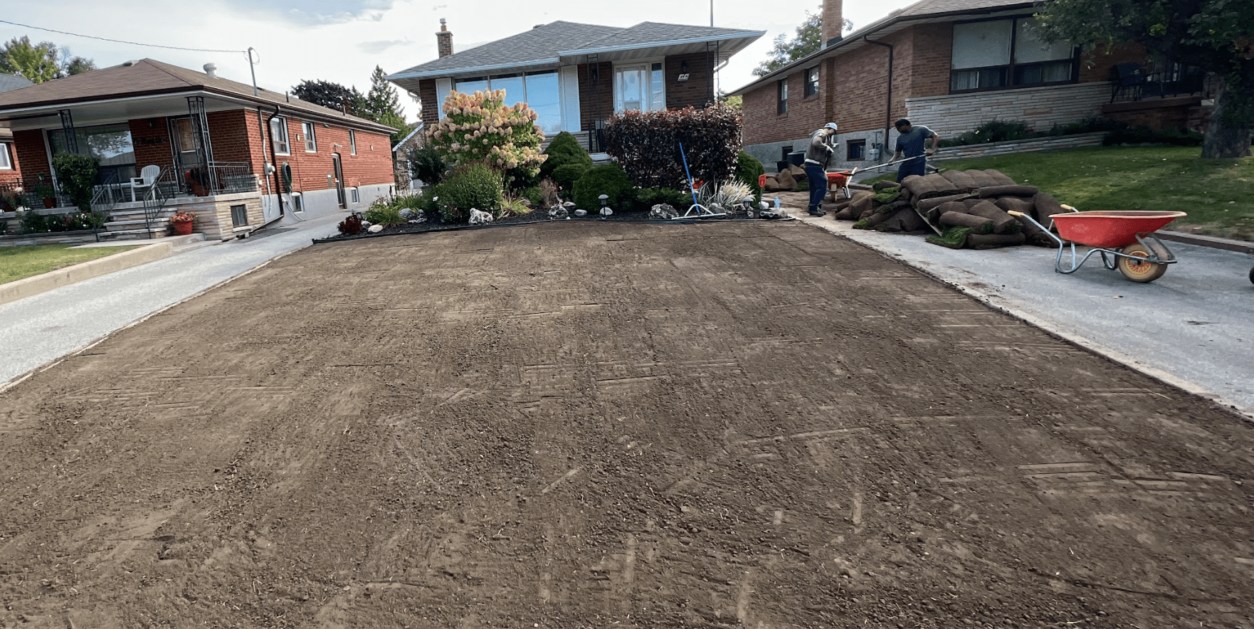 People lay sod on a front yard, with brick houses in the background and gardening tools scattered around.